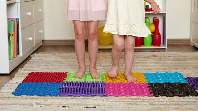 Two Children Are Standing On The Spot, Standing On Orthopedic Massage Mat With A Surface Of Different Texture And Hardness. Prevention Of Flat Feet. Kids In The Children's Room Folded A Mat Of Puzzles