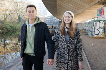 Portrait happy young couple holding hands, walking along canal