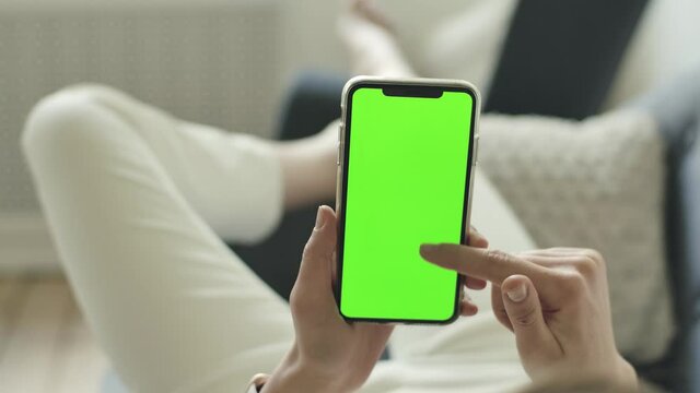 Young adult female person browses smartphone in hands. Empty chromakey display, mock up space for advertisement logos, texts. Woman lies on sofa, scrolls green mobile phone screen for ads, adverts.