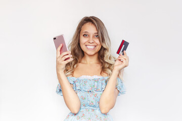 A young charming smiling blonde woman with wavy hair holds a plastic credit card and mobile phone in her hands to pay for purchases in the internet isolated on a white background. Shopping online