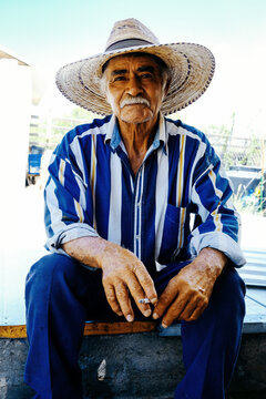 Portrait of smiling senior male farmer wearing hat and striped shirt holding cigarette while sitting on bench