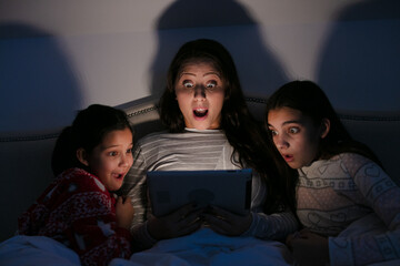 Surprised mother and daughters watching movie on digital tablet in dark bedroom