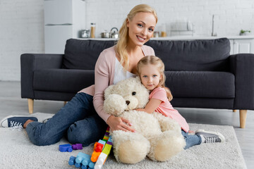 mother and daughter embracing on floor near teddy bear and colorful building blocks