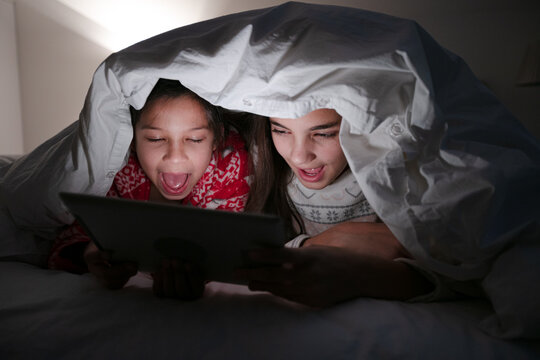 Sisters Watching Movie On Digital Tablet Under Blanket In Dark Bedroom