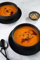 Two deep black bowls with an orange pumpkin cream soup with pumpkin seed oil, two black spoons on a marble surface