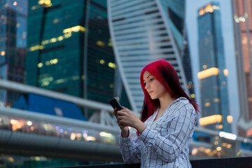Beautiful red-haired girl with smartphone in the evening on lighted city street.