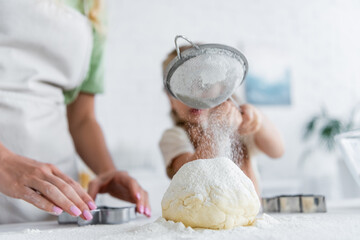 blurred girl sifting flour on dough near mom in kitchen