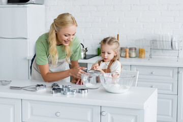Fototapeta na wymiar girl sifting flour with sieve into bowl near happy mother