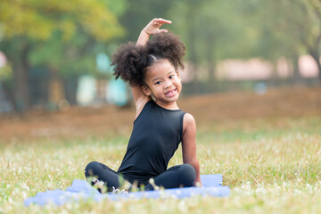 Smiling African American little girl practicing yoga and stretching her hands on exercise mat outdoor in the park