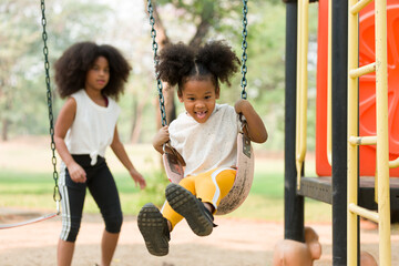 Happy cheerful African American child girl playing on swing at playground in park. Smiling girl having fun on a swing