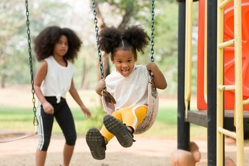 Happy cheerful African American child girl playing on swing at playground in park. Smiling girl having fun on a swing - Powered by Adobe