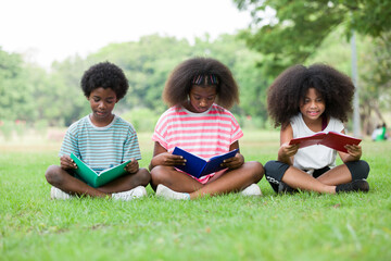 Children reading book on grass outdoor. Group of African American children in casual wear reading book while sitting on green grass in the park, against green summer garden