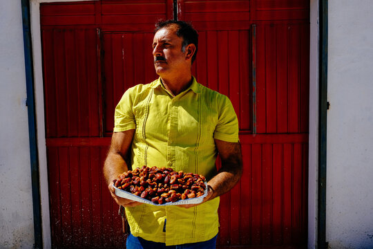 Mature male farm worker looking away while holding fresh dates in foil container against closed door on sunny day