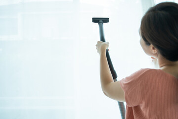 Asian young woman using vacuum machine to cleanup the dust on curtain in living room close up. Housework and chores in daily life concept. Housekeeper cleaning a white curtain.