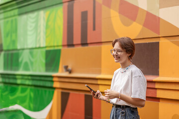 a teenage girl, brunette, with a mobile phone, against the background of a bright wall with graffiti on a warm sunny day