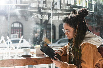 pretty young white hispanic latin girl with dreadlocks and glasses using her tablet at the bar of a coffee shop next to the display case.