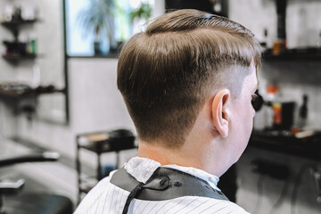 Visit to the barbershop. Hairdresser, barber shaves client's temples,sides with hair clipper. Young stylish man makes fashionable haircut.Customer sitting in dark men's beauty salon covered with cape