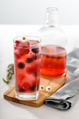Tall glass with red fruit juice on ice and bottle in the foreground with small flowers