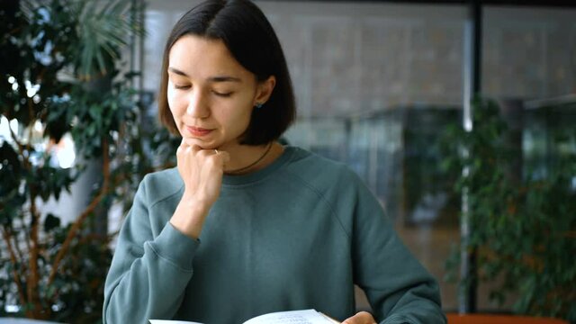Close-up portrait of a young cheerful female student, she is looking for the necessary information in a book and writes in a notebook, preparing for a lesson. Education concept.