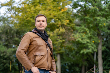 Obraz premium Outdoors portrait of smiling handsome millennial man with backpack on one shoulder standing at autumn park, looking at camera