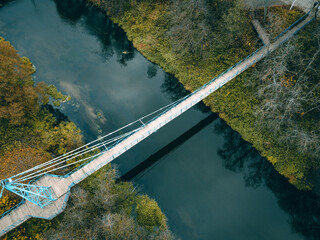 Fototapeta premium a small cable-stayed bluish-white bridge with a wooden floor over a narrow river, green grass and bushes on the sides