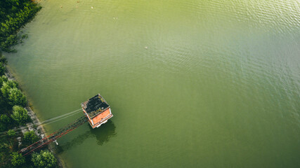 Obraz premium a marsh-colored lake, in the corner there are green bushes on concrete slabs, a small red-brick building with a black roof stands on the water across a small iron bridge