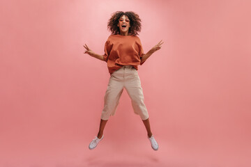 full-length image of happy African woman jumping against background of wall of pastel pink color. young brunette in sunglasses with curly hair shows peace sign with two hands.