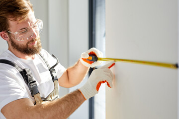Man repairman builder in overalls and gloves, safety glasses measures white wall with tape tool,...