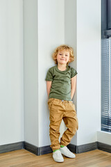 Adorable little blond kid boy standing near window in corner and looking at camera smiling, indoors. Caucasian child in casual outfit at home, alone. portrait, side view