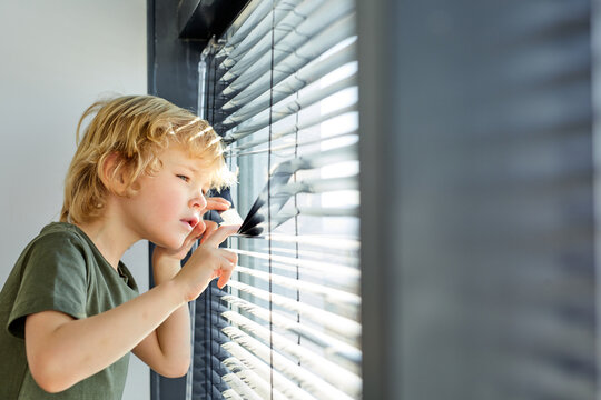 offended little caucasian boy looks out the window, lowers the blinds, spies, boy is left at home, forbidden to go out, boy is punished. side view portrait