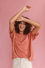 Obraz na płótnie Canvas Cheerful dark-skinned girl laughs strongly raising her hands up while standing on pink background. Curly brown-haired woman with good mood is dressed in casual clothes. Beauty and youth concept
