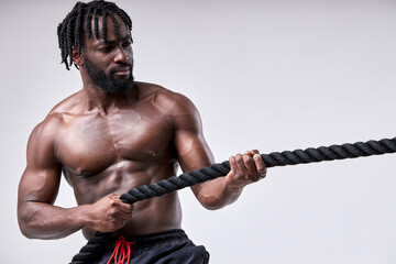 handsome young fit bearded afro sportsman pulling rope strongly isolated on white studio background. Black shirtless guy engaged in cross fit. Concept of sport, bodybuilding, healthy lifestyle.