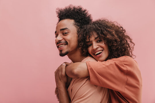 close-up studio shot of two charming partners on pink background. smiling curly brunette hugs bearded guy from behind with two hands.