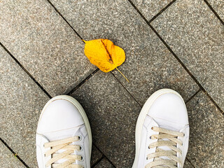 A person in white lather sneakers standing in front of a yellow autumn fall leaf in a city park