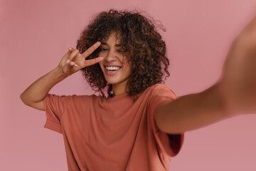 cute radiant lady makes selfie showing two fingers near her face on pink background. dark-haired, curly, smiling African woman with snow-white smile blinks one eye.