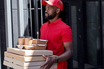 Young bearded african delivery man in red uniform with pizza boxes, lunch box and coffee, looking at side confidently, ready to serve customers clients. Side view. Delivery service, takeaway orders