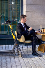 Young guy sit outdoors in cafe behind table using laptop, working online, side view. Guy in black suit sitting alone, using electro scooter, modern technologies concept. business people, lifestyle