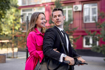 married couple in stylish bright clothes driving electric scooter on city street, side view portrait of beautiful man and woman in suit and dress having fun, enjoying walk on scooter, have rest