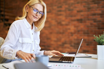 Attractive mature businesswoman in glasses using laptop and doing some paperwork at the office