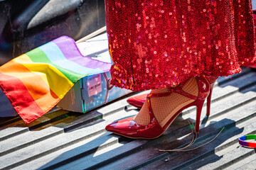 Close up of a drag queen's red stiletto heels, sparkly dress on a pickup truck's bed, with rainbow...