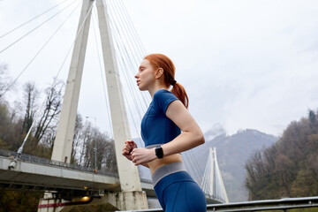 View from down on confident female runner jogger in motion, looking at side, wearing blue sportive clothes, red-haired lady on modern bridge. copy space. people lifestyle, sport, workout concept