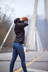 Rear view on young female in sportswear walking on bridge having rest after running workout, at cold spring day, redhead slim female outdoors, healthy active lifestyle, sport and fitness concept