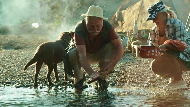 Senior Couple Camping Is Washing Dishes In A Stream