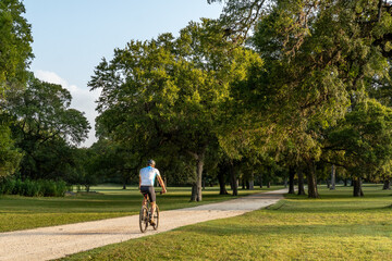 Man, person, rider on a bicycle along a path through a grove of oak trees and grass level area, Swede Creek Park, Cordillera Ranch, Boerne, Hill Country, Texas