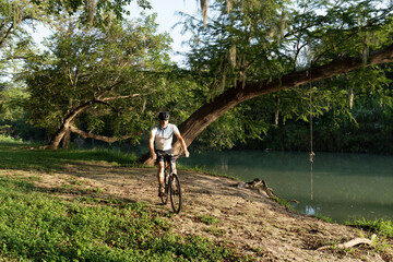 Man, person, rider on a bicycle along a river with bent trunks of oak trees and rope swing over the...