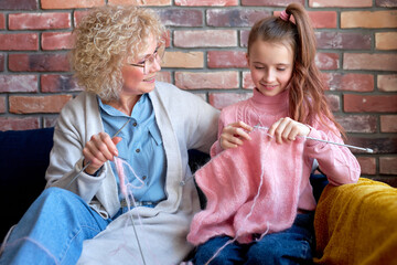 Smiling loving senior grandmother is teaching little cute daughter knitting, sitting on cozy couch...