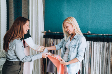 Young woman designer talking with a saleswoman and chooses fabrics for curtains in textile show...
