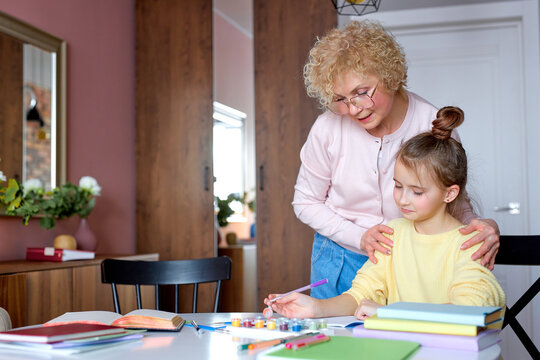 pretty girl and grandmother sit behind desk at home, girl is drawing. Cute child and senior woman having fun together. Happy family indoors. Aged grandma support daughter, sit next to girl