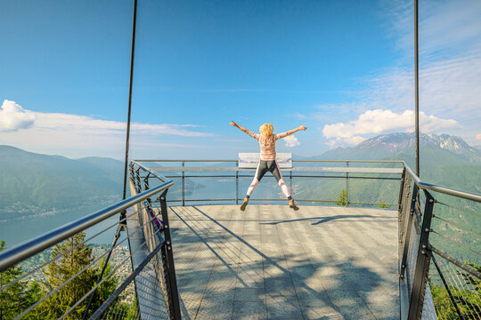 Woman jumping on Aussichtsplattform platform of Cardada-Cimetta mount in Switzerland. Skyline of Locarno on Cardada mount. Lake Langensee cityscape in Ticino.