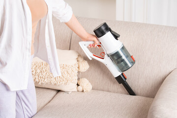 A woman is cleaning the sofa with a cordless vacuum cleaner. House cleaning. - obrazy, fototapety, plakaty
